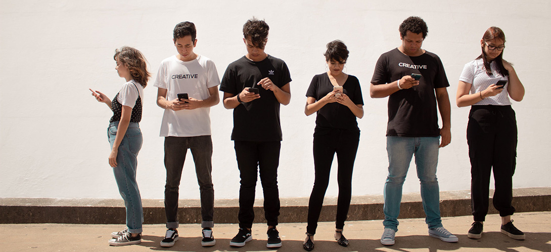 A group of teenagers standing in a line all on their mobile phones.