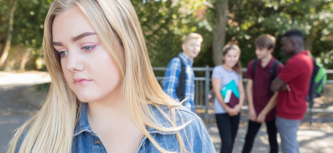 A close up image of a girl standing in the foreground to the left looking downwards while a group of her friends are blurred out whispering in the background.