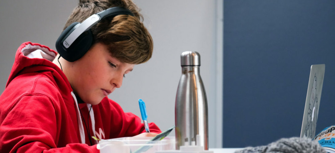 A young boy sitting at a table with an open laptop, writting while listening to headphones.