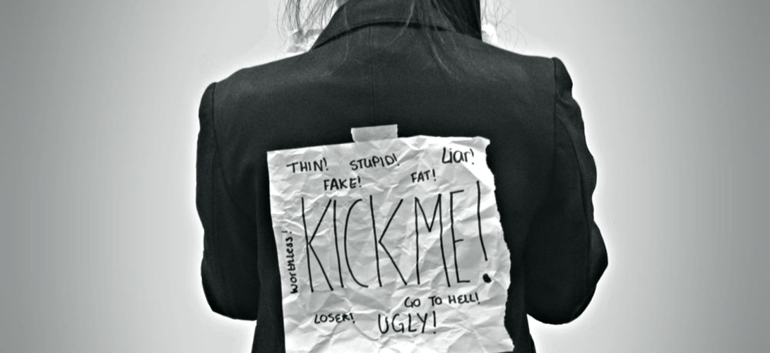 A teenager with a piece of paper stuck on their back with the words "Kick Me!" in larger letters surrounded by the words "Thin, stupid, liar, fake, fat, worthless, loser, ugly and go to hell".