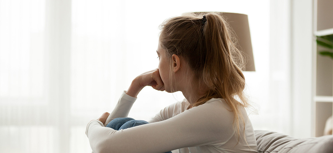 A teenage girl sitting on a sofa clutching her knees into her body. She is resting her elbow on a knee with her head on her hand looking out towards a window with net curtains pulled closed.