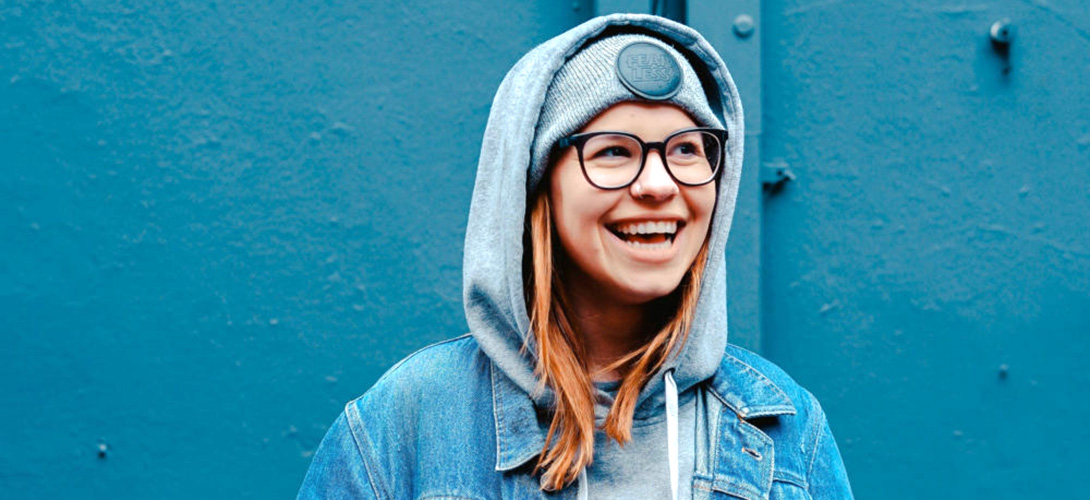Girl smiling wearing a hoody, denim jacket and glasses standing against a blue background.