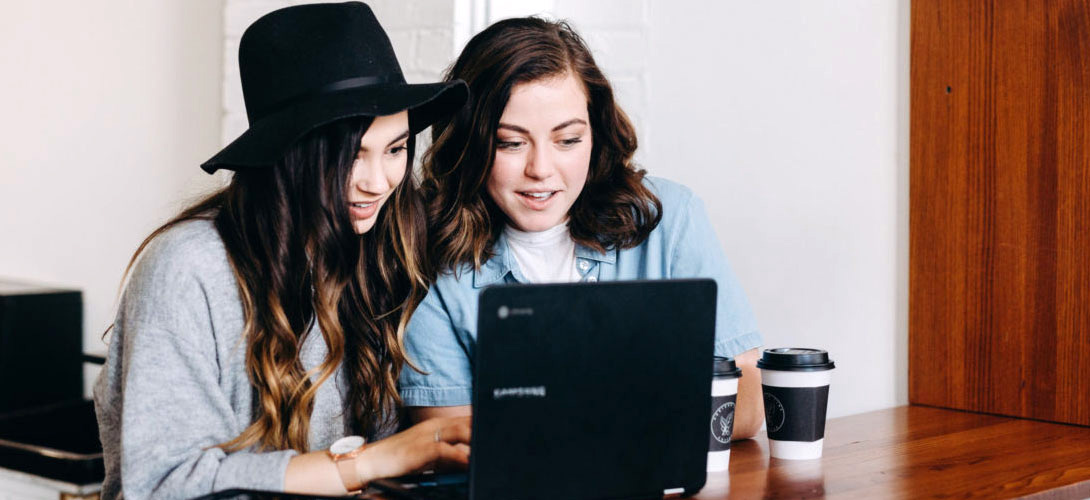 Two teenage girls sitting at a table laughing looking a laptop.