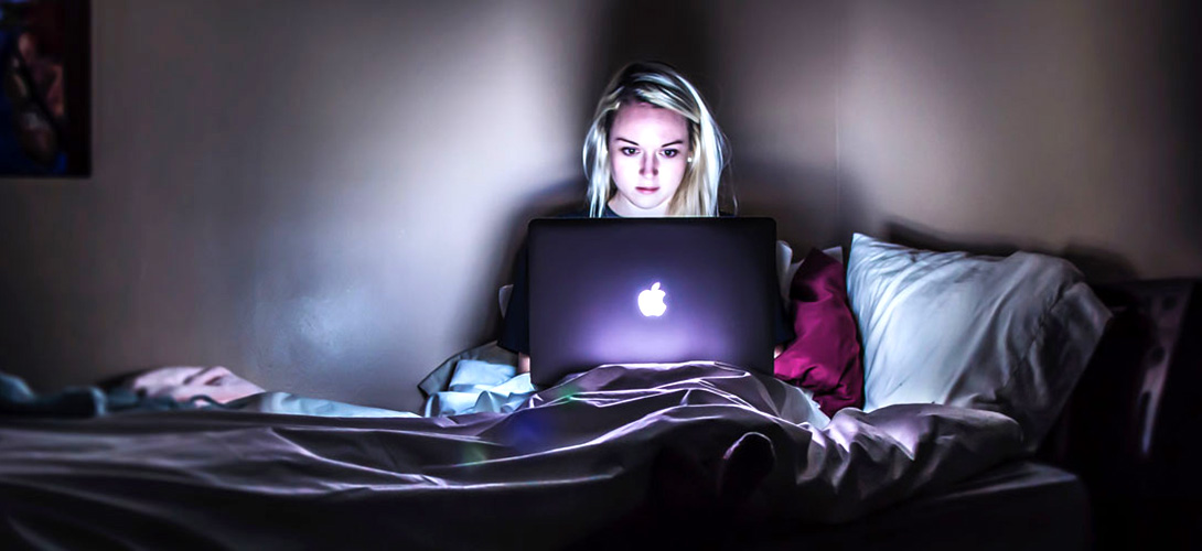 A teenage girl sitting on her bed alone in the dark looking at her laptop screen.