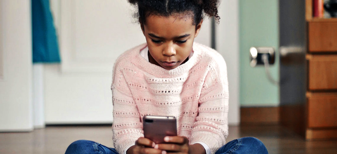 Young girl sitting alone in her home looking down at her mobile phone with a sad and serious expression on her face.