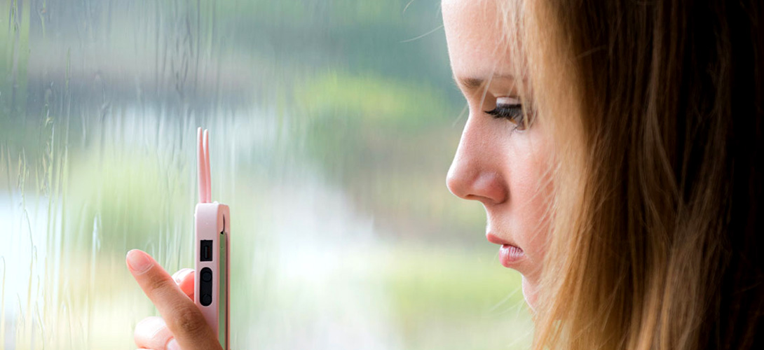 A young teenage girl standing in front of a window looking at her mbile phone with a sad expression on her face.
