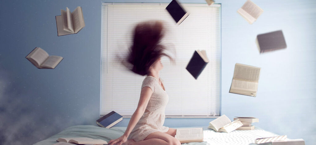 A teenage girl sitting on her bed throwing her head back with her books tossed in the air around her.