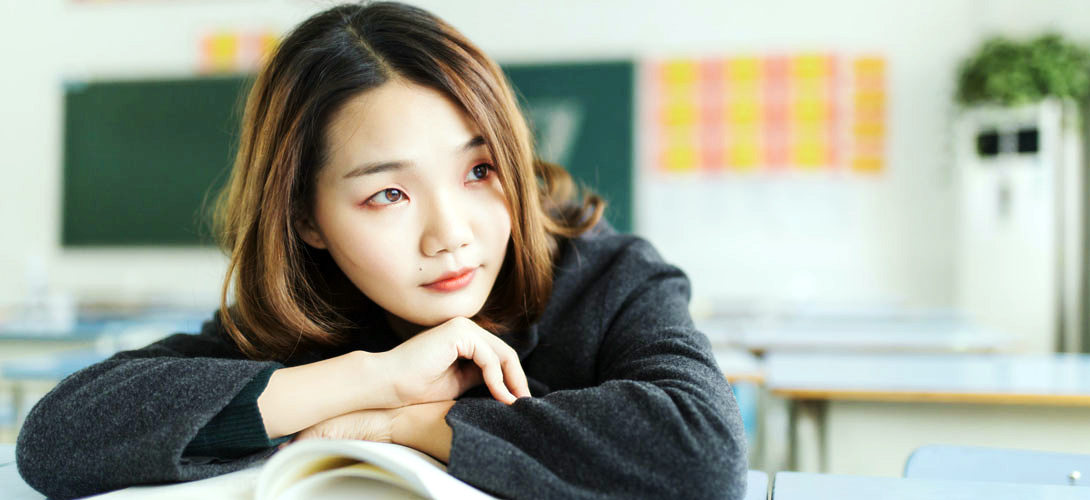 A teenage girl sitting on her own in an empty classroom with her arms folded across an open book.