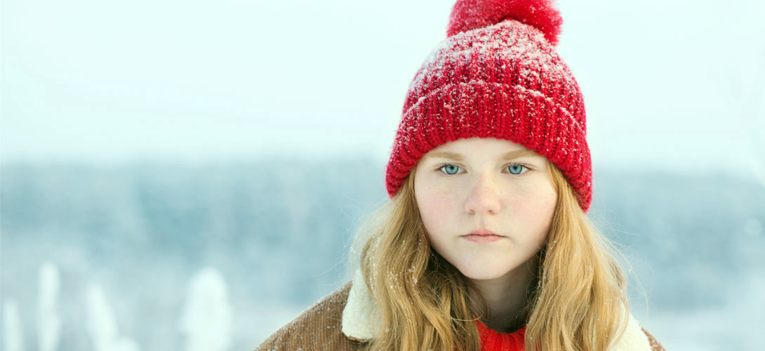 Young teenage girl wearing a red wool hat dusted with snow. A snow scene blurred in the background. The girl looks sad.