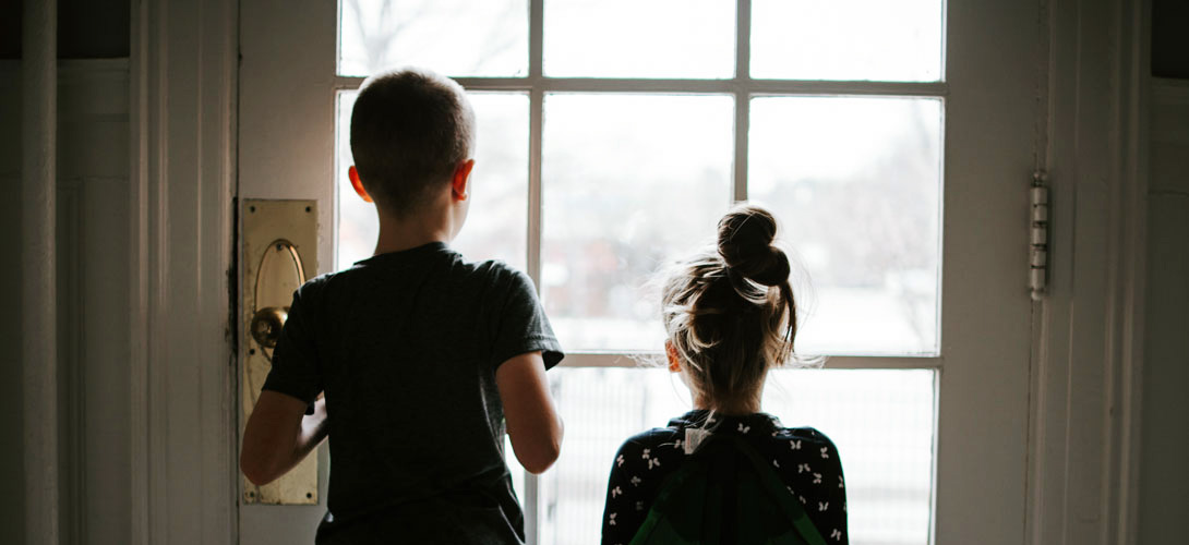 An older brother and younger sister stand side by side looking out a glass door.