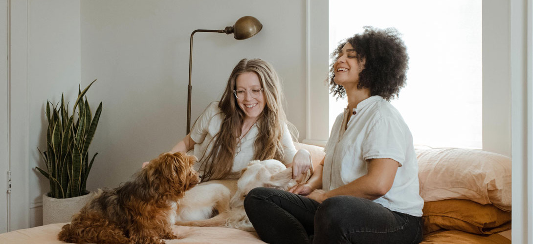 Two friends chatting and smiling sitting with a dog on a bed.