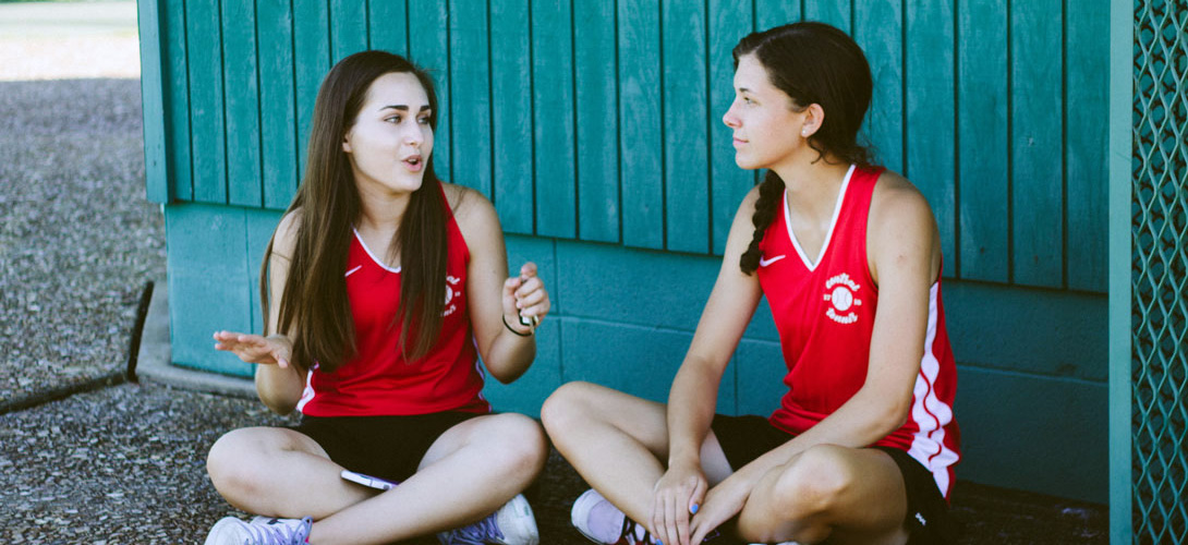 Two teenage girls sitting at a sports ground chatting.