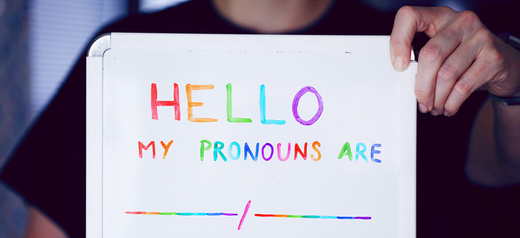 A person holding up a notice board with the words "Hello my pronouns are ________ / _______" spelt out with different coloured markers.