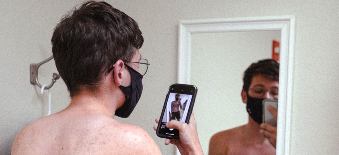 A young boy wearing a black facemask standing in front of a mirror taking a selfie.