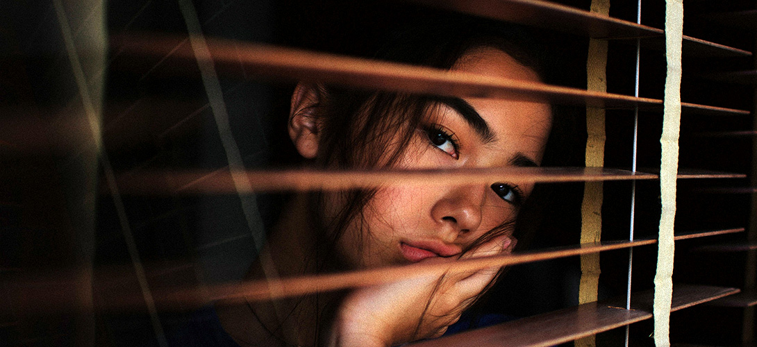 A young teenage girl with a bored expression looking out a window through the gap in wooden blinds.