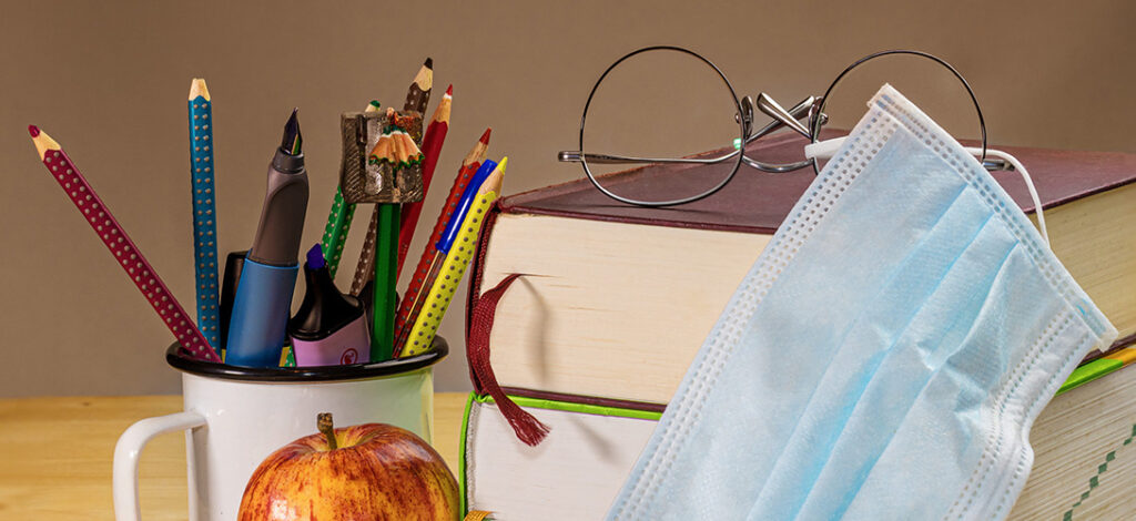 Mug with pencils and highlighters, an apple, school books with a pair of glasses on top and a surgical mask lying against them.