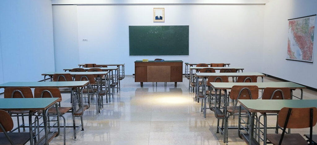 An empty classroom with pupils desks, chairs, a teacher's desk and chair and a blackboard.