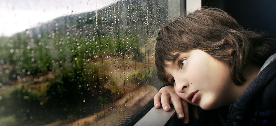 A boy with a sad expression resting his head against a window looking out to the rain.