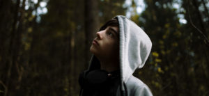 A young boy standing against a background of trees, wearing a grey hoodie with the hood pulled up. He is looking up into the sky.