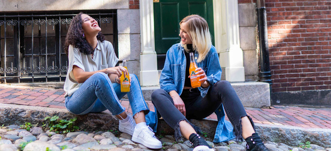 Two young women sitting a curb side looking at each other and laughing and smiling while drinking a bottle or soft drink.