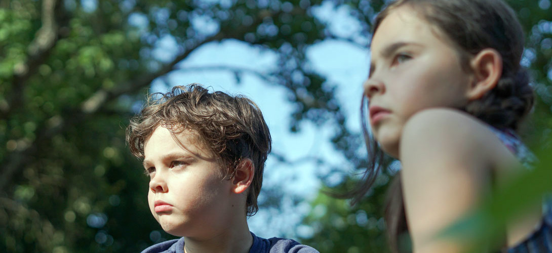 A close-up of a young boy and girl sitting against a background of trees nad leaves, looking into the distance not communicating.