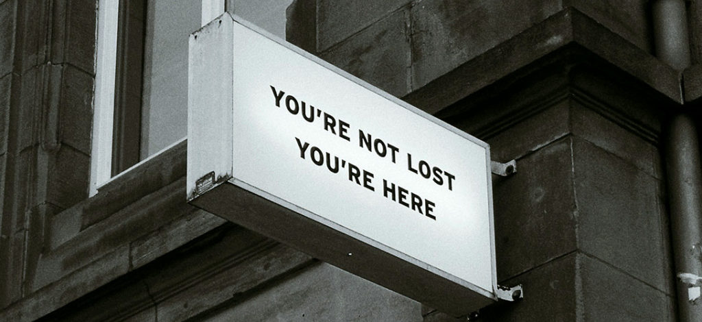 A white sign with black writting on the side of a building saying you're not lost you're here.