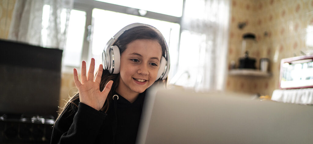 A girl wearing headphones smiling and waving at her laptop screen.