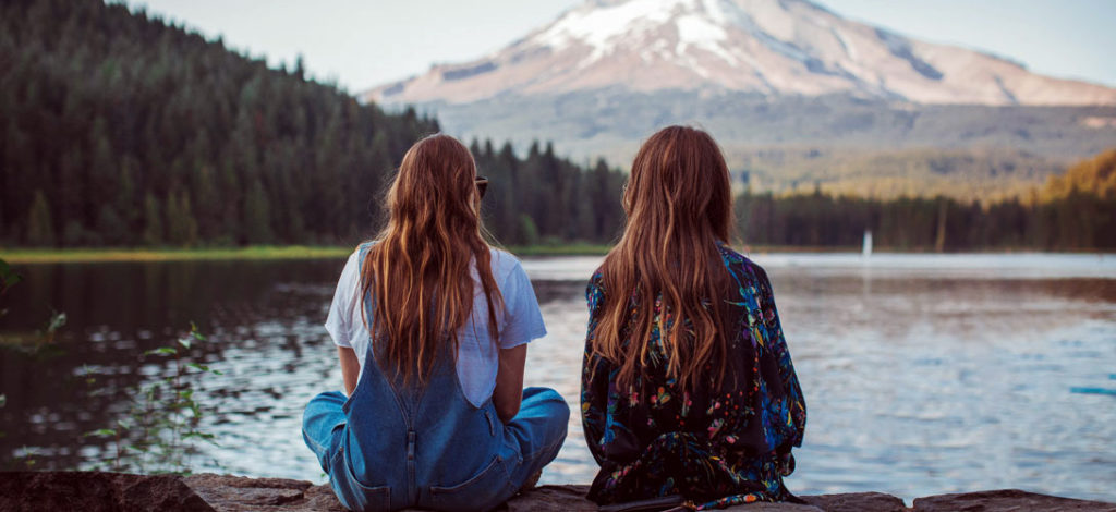 Two teenage girls sitting by the side of a lake or river facing in the direction of mountains in the distance.