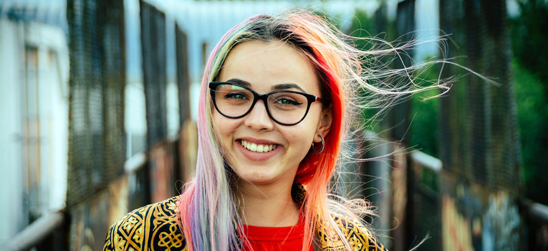 A young girl with colourful hair and wearing glasses looking straight ahead and smiling.