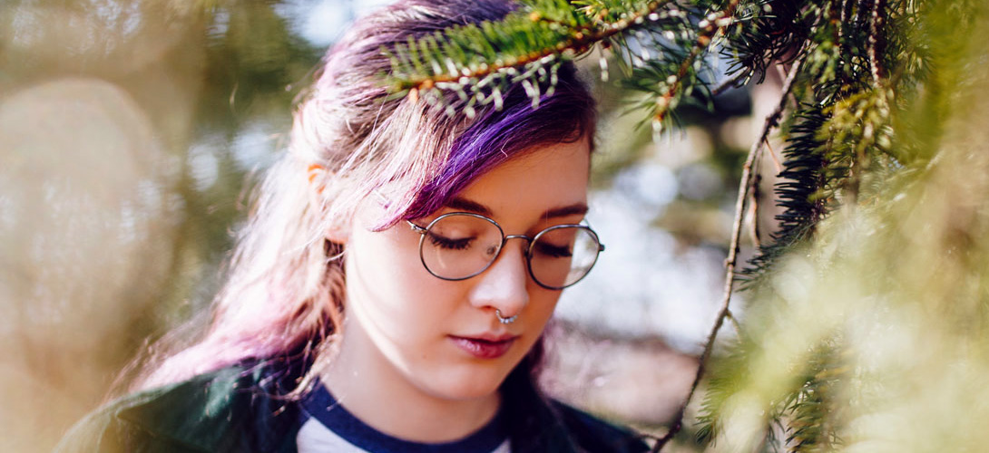 Close-up of a girl wearing glasses with purple streaks in her hair. She has a serious and sad look on her face, she is standing beside trees and looking down at the ground.