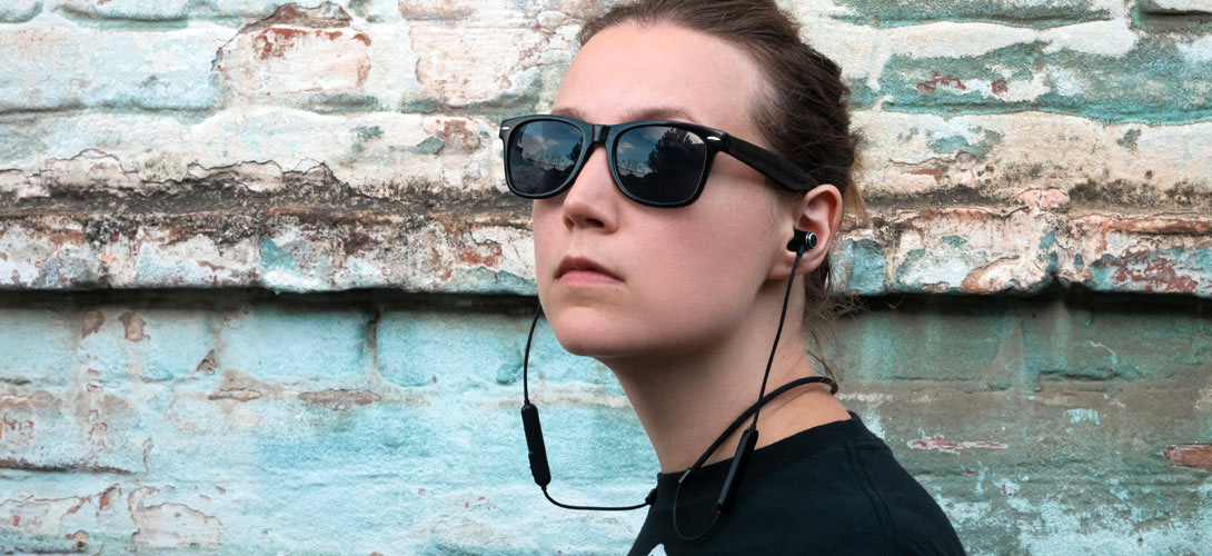 A teenage girl standing in front of a blue and white brick wall. She is wearing sunglasses and earpods.
