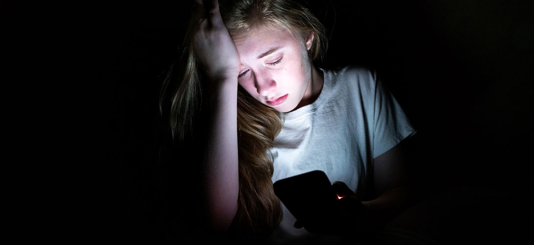 A teenage girl is sitting in the dark upset as she looks down at her phone with her head resting against her hand.
