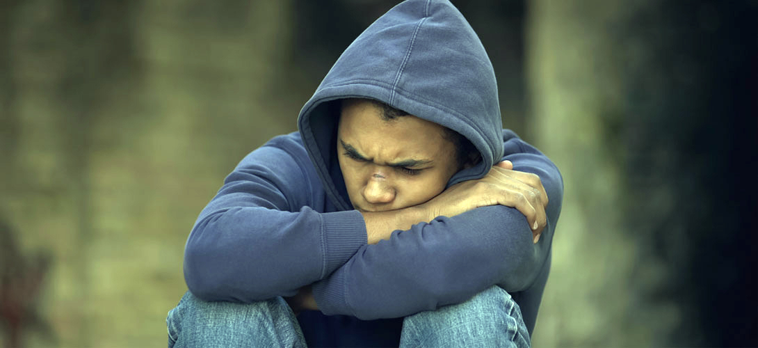 A teenage boy in a hoodie sits with his arms folded leaning his head in his arms and looks down with puzzled look on his face.