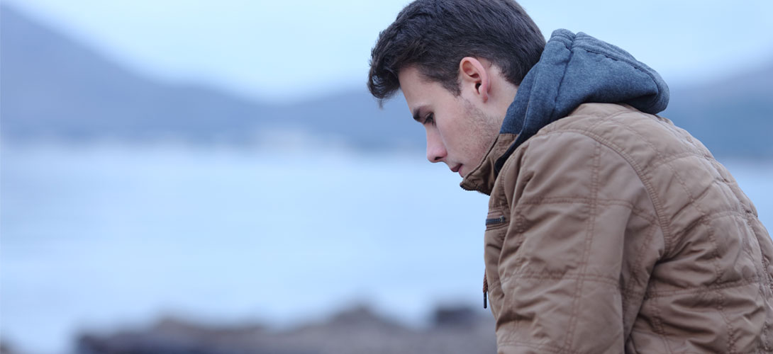 An older teenage boy is sitting at the side of a lake or the sea wearing a jacket and hoodie. He has a sad expression and is looking down at the ground.