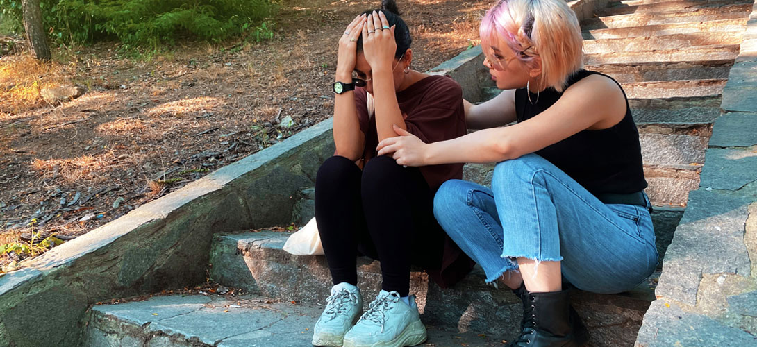 Two teenage girls sitting on steps. The girl on the left has her hand covering her head. The girl sitting next to her has her arms around her comforting her.