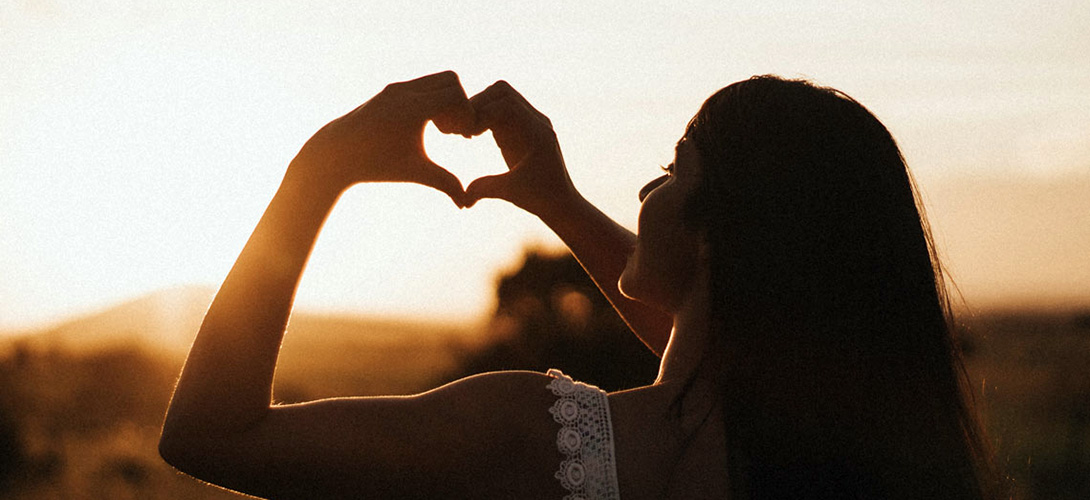A teenage girl looking into the sunset and making a heart shape with the fingers of both hands.