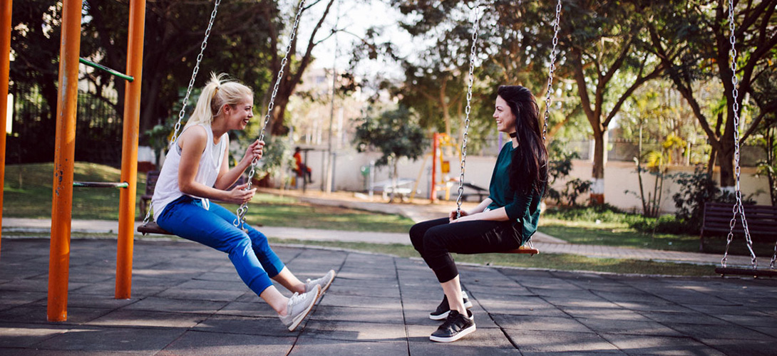 Two teenage girls sitting on swings facing each other, laughing, smiling and chatting,