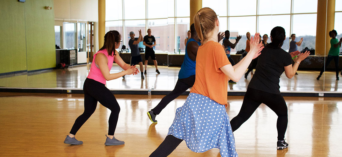 Group of young children taking part in a dance class facing a mirrored wall while stepping and clapping their hands.