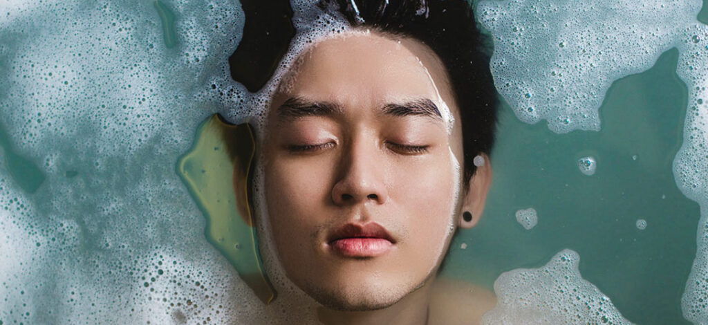 A close-up of a teenage boy face, lying in a bath with his eyes closed and his hair submerged in water,