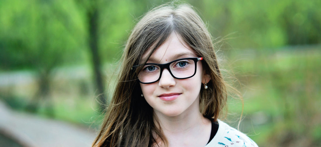 A close-up of a young girl with long brown hair and wearing glasses looking straight ahead and smiling.