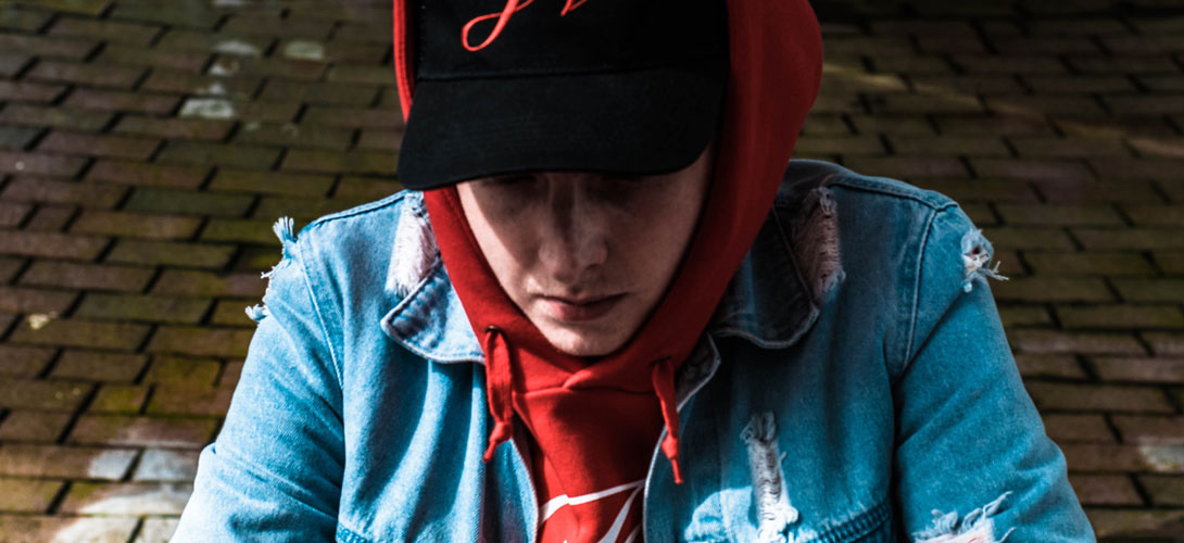 A close-up of a teenage boy wearing a cap with a hoodie over it, sitting on the ground, his head bent down looking at the ground.