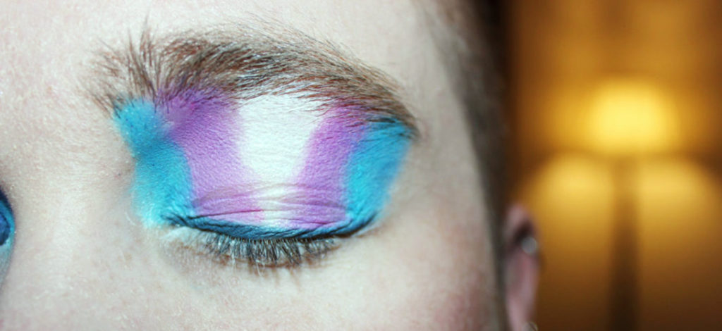 A close-up of a teenagers closed left eye with vertical stripes of coloured eyeshadow blue, purple, white, purple and blue.