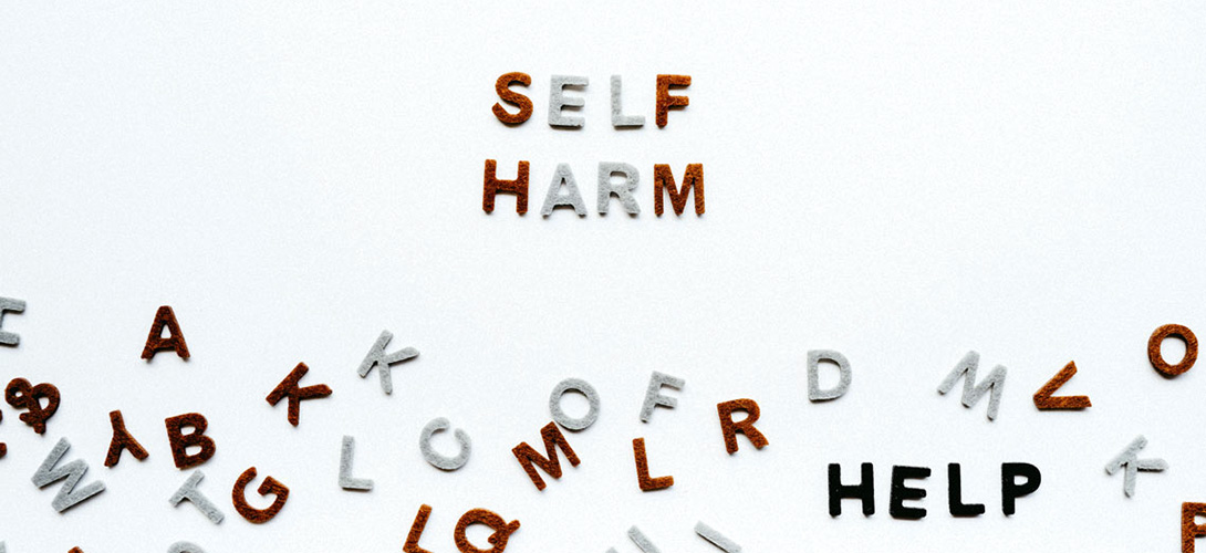 A white background with the words self harm spelt out in red and grey letters. Other letters are lying across the bottom of the image with the word help spelt out in black letters.