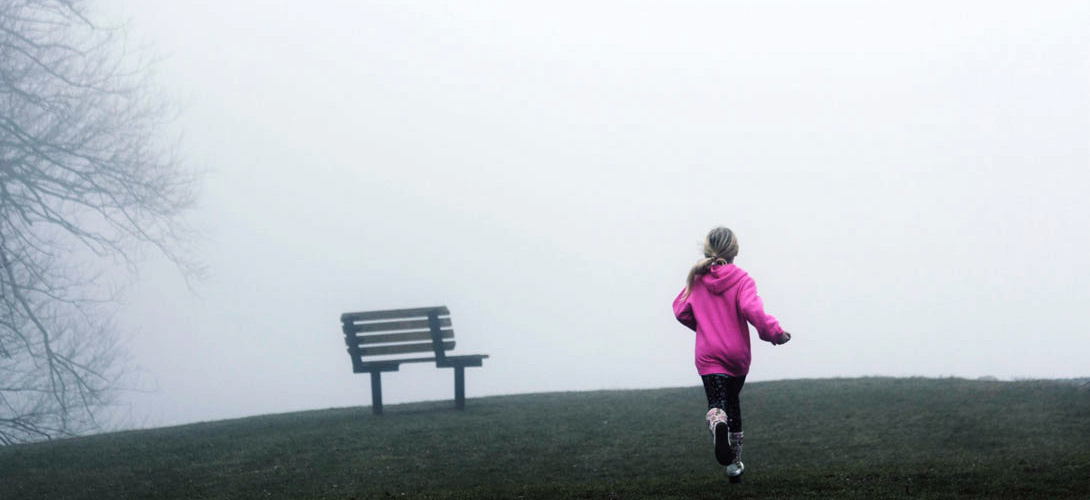 A misty day with girl running across some grass to a bench.