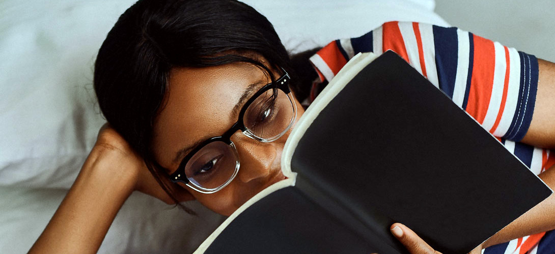A teenage girl lying on her bed supporting her head with one hand and holding her book with the other while she is reading.